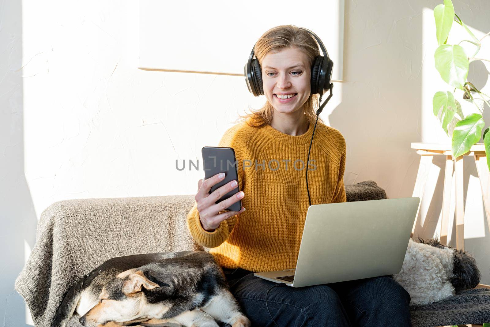 Distance learning. E-learning. Young smiling woman in yellow sweater and black headphones studying online using laptop, sitting on the couch at home