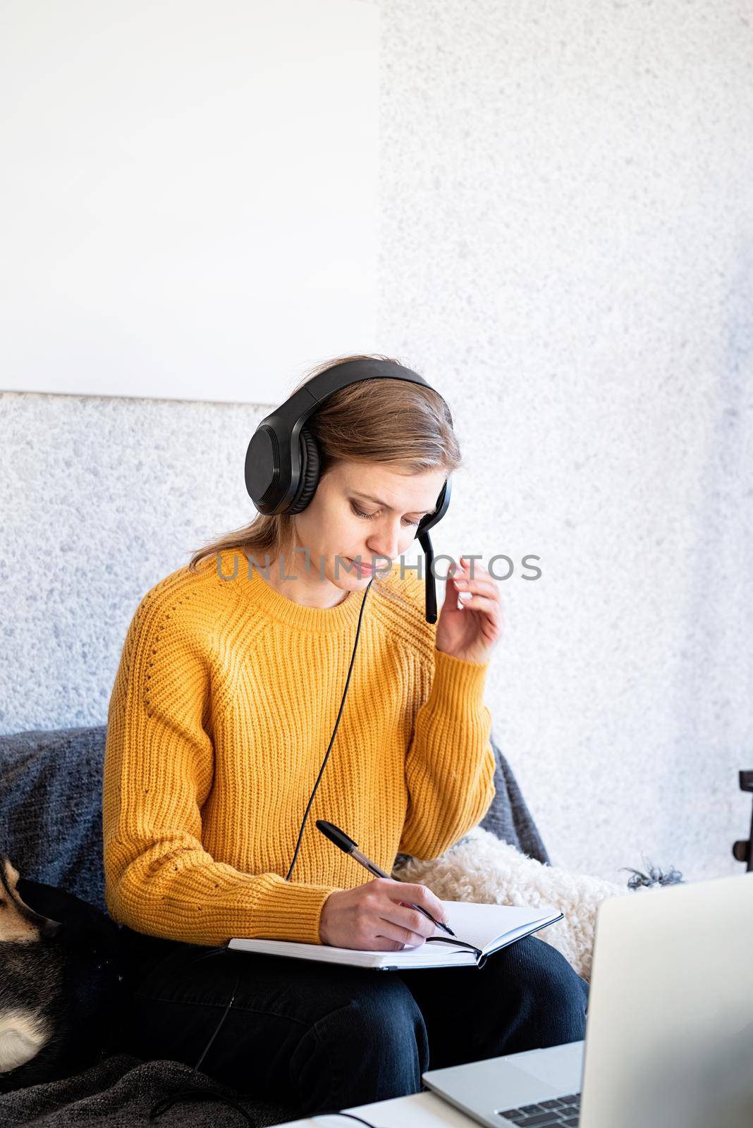 Distance learning. E-learning. Young smiling woman in yellow sweater and black headphones studying online using laptop, sitting on the couch at home