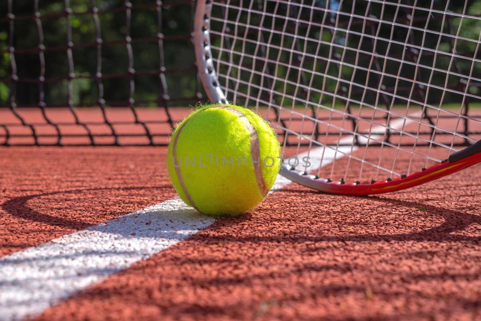 Tennis ball, line and racket on an outdoor court with the racket standing on end casting a shadow across the all weather surface