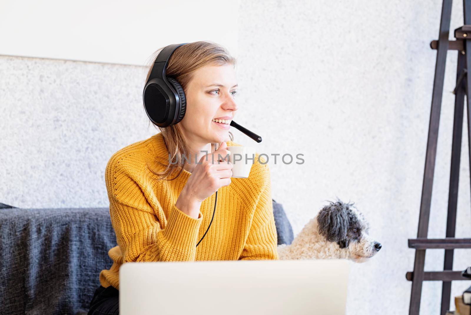 Distance learning. E-learning. Young smiling woman in yellow sweater and black headphones studying online using laptop, sitting on the couch at home