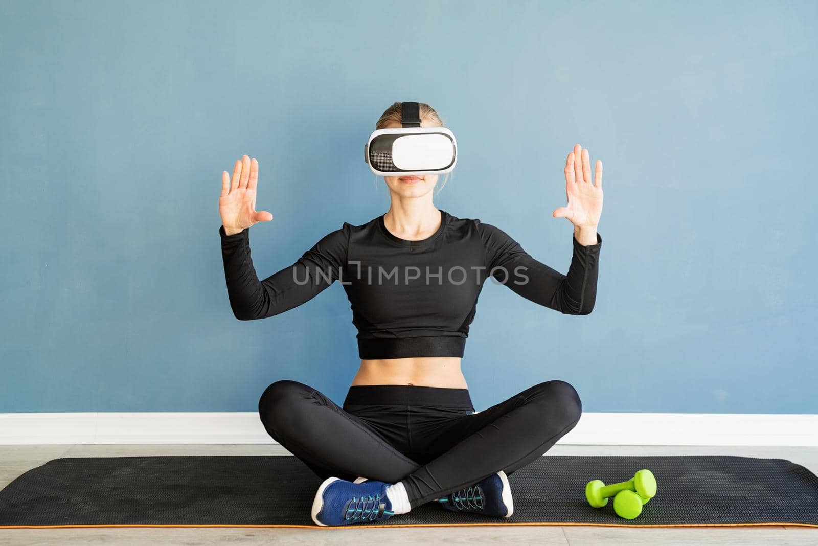 Fitness, sport and technology. Young athletic woman wearing virtual reality glasses sitting at fitness mat using VR interactive menu at blue background