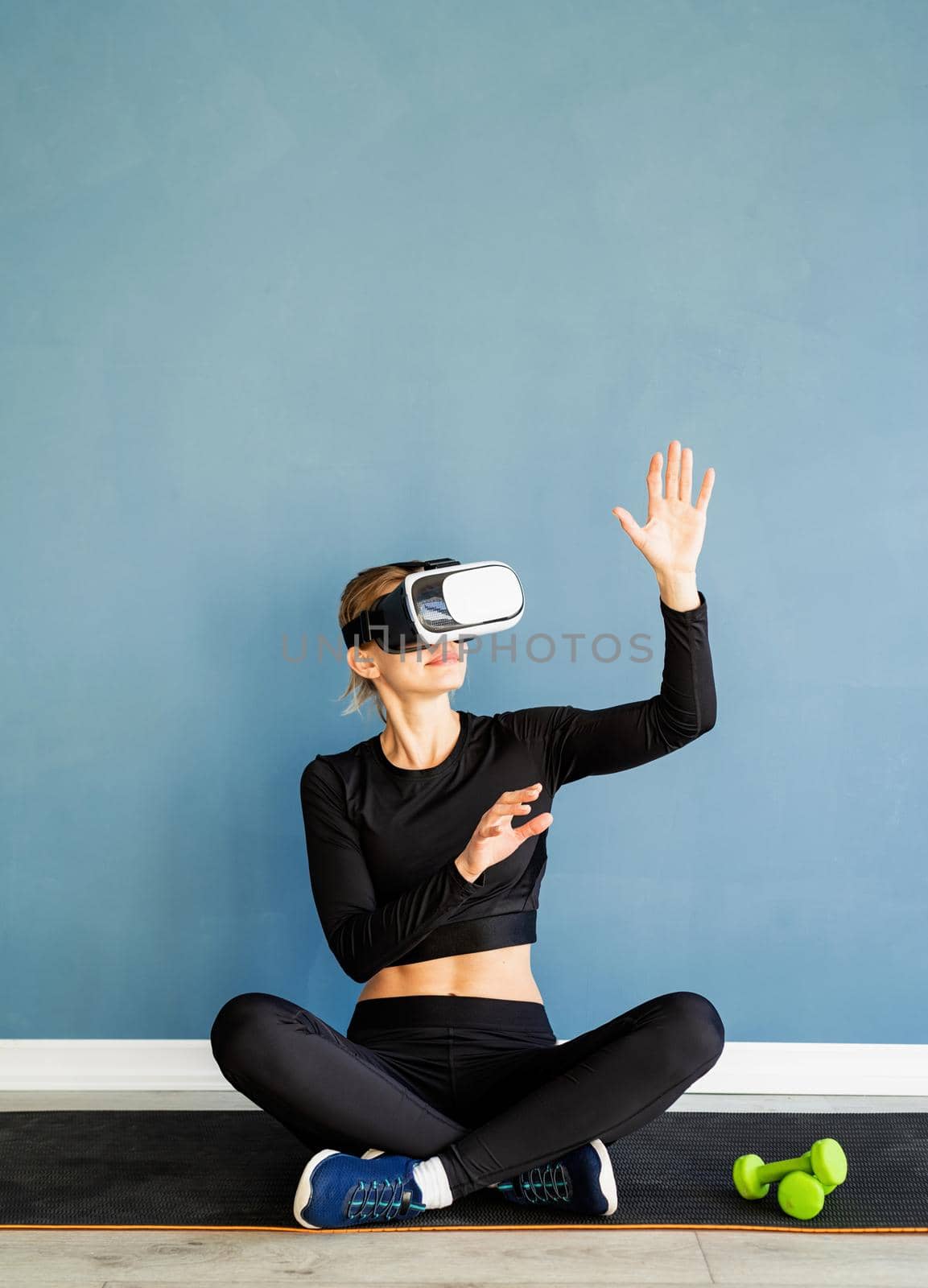 Young blonde woman in sport clothes wearing virtual reality glasses sitting at fitness mat using VR interactive menu by Desperada