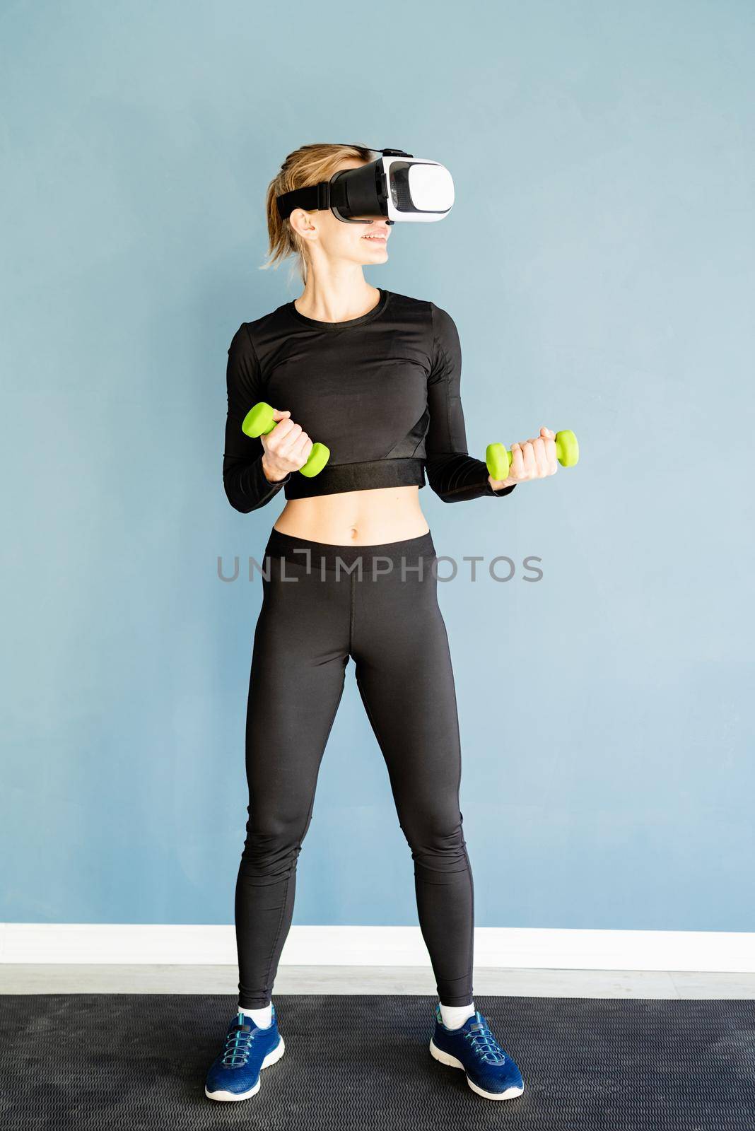 Fitness, sport and technology. Young athletic woman wearing virtual reality glasses standing at fitness mat working out with dubbells