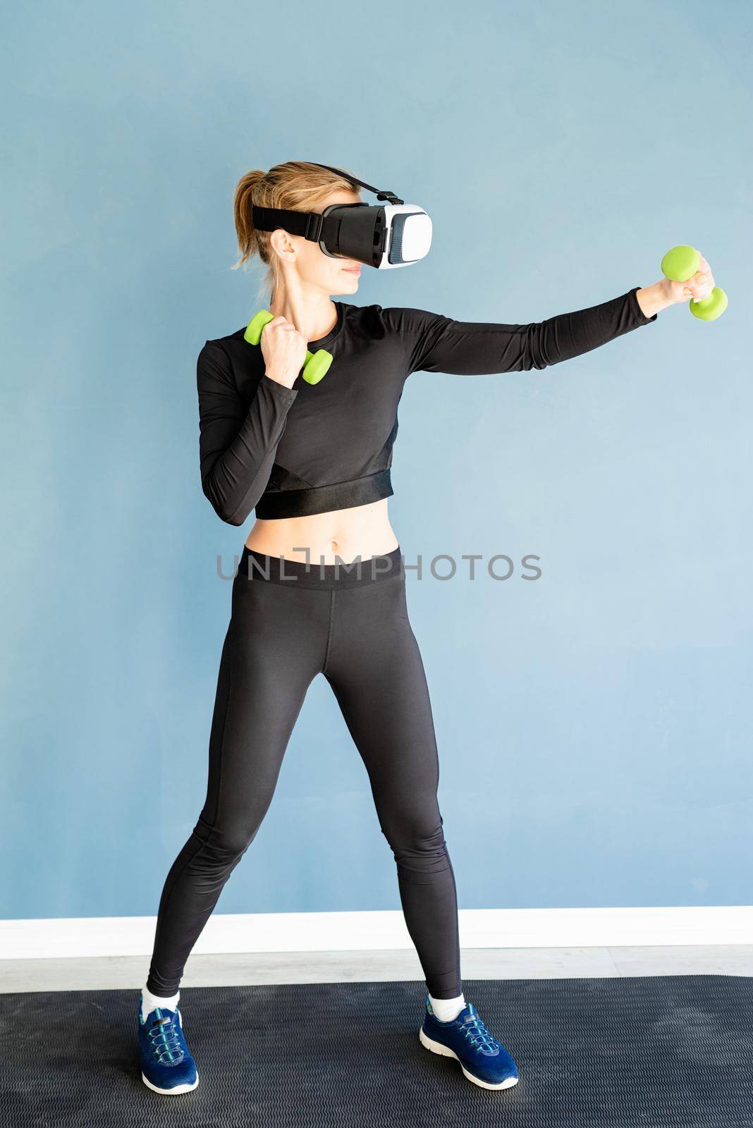 Fitness, sport and technology. Young athletic woman wearing virtual reality glasses standing at fitness mat working out with dubbells