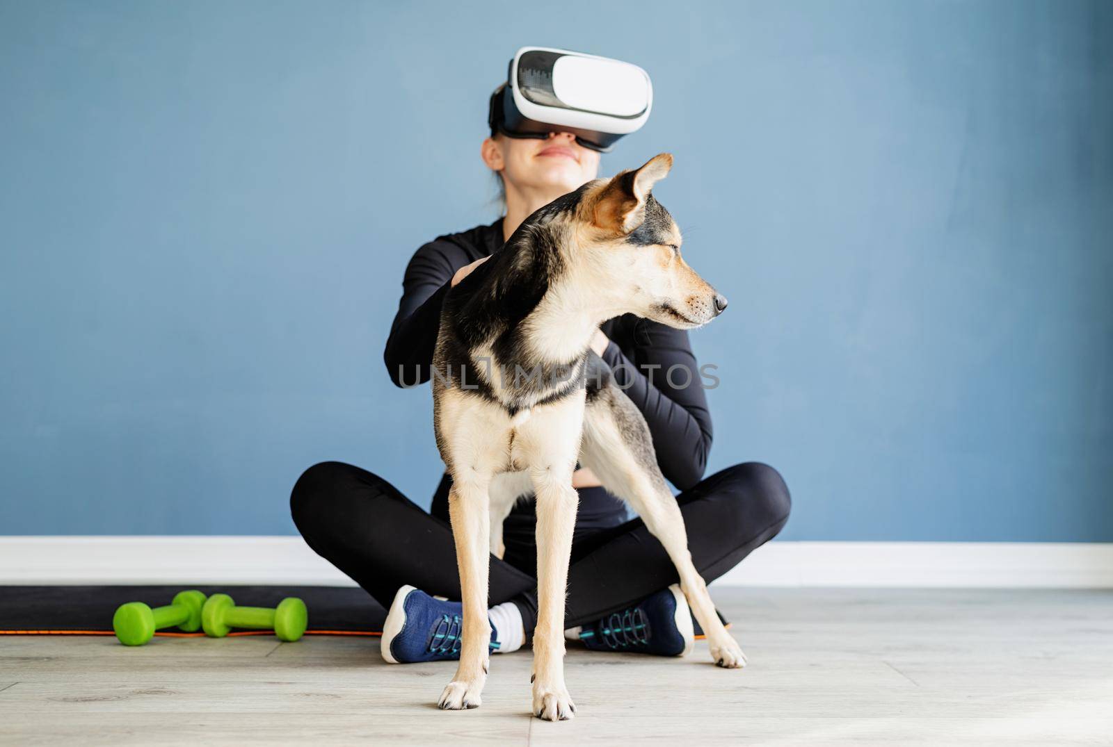 Fitness, sport and technology. Young athletic woman wearing virtual reality glasses sitting on fitness mat with dog