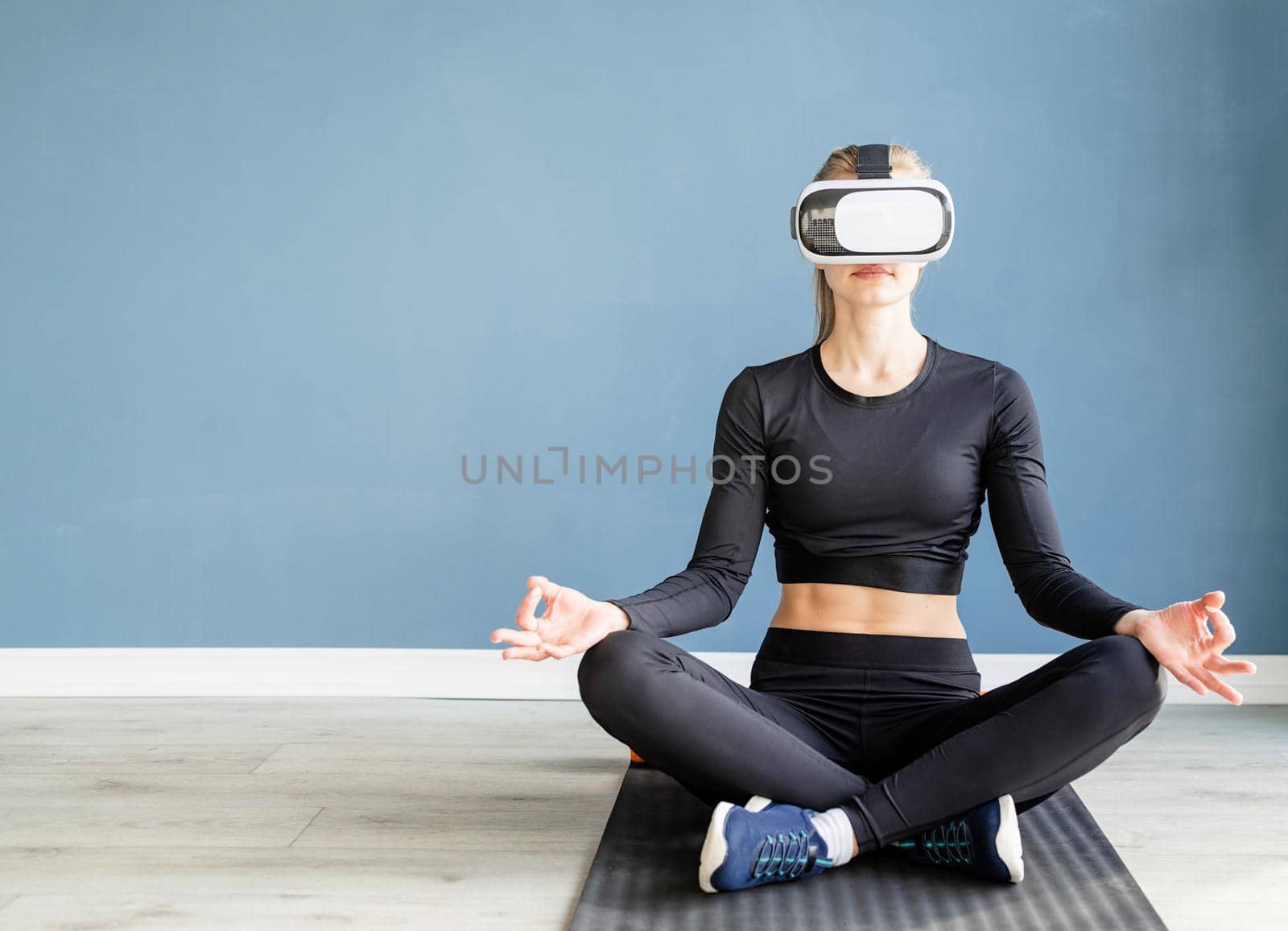 Young blonde woman in sport clothes wearing virtual reality glasses meditating on fitness mat by Desperada