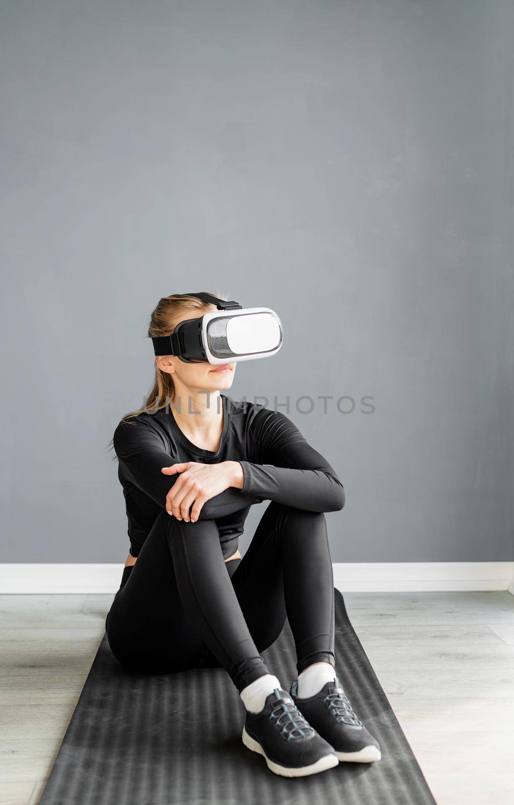 Young blonde woman in sport clothes wearing virtual reality glasses sitting on fitness mat by Desperada