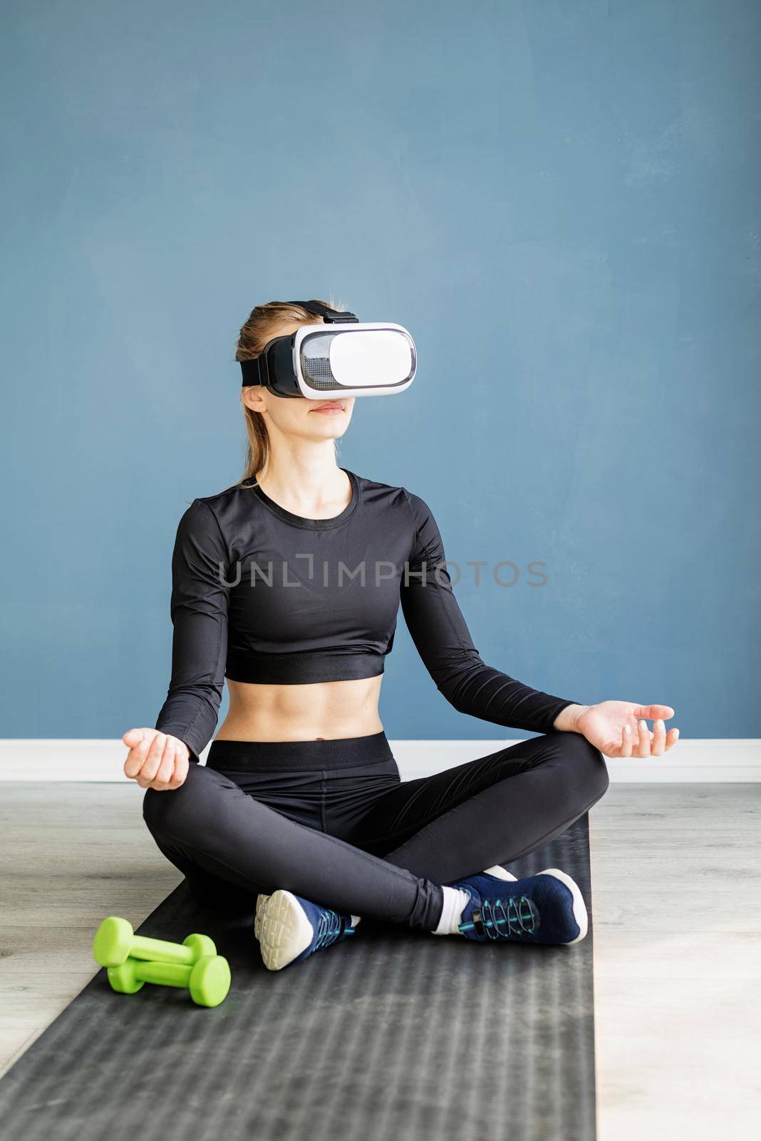 Young blonde woman in sport clothes wearing virtual reality glasses meditating on fitness mat by Desperada