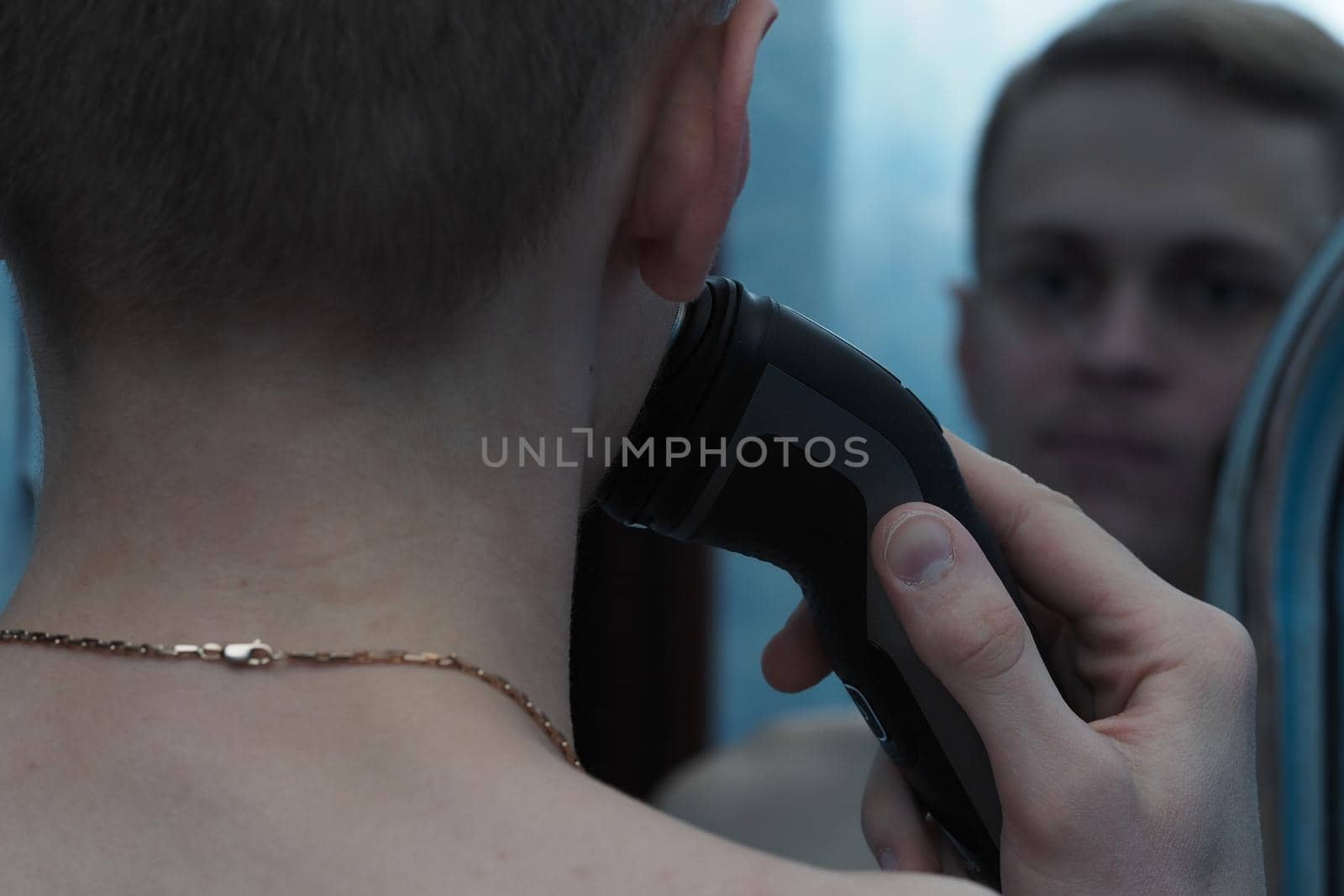 A young man of European appearance shaves his face with an electric razor and looks in the mirror. High quality photo