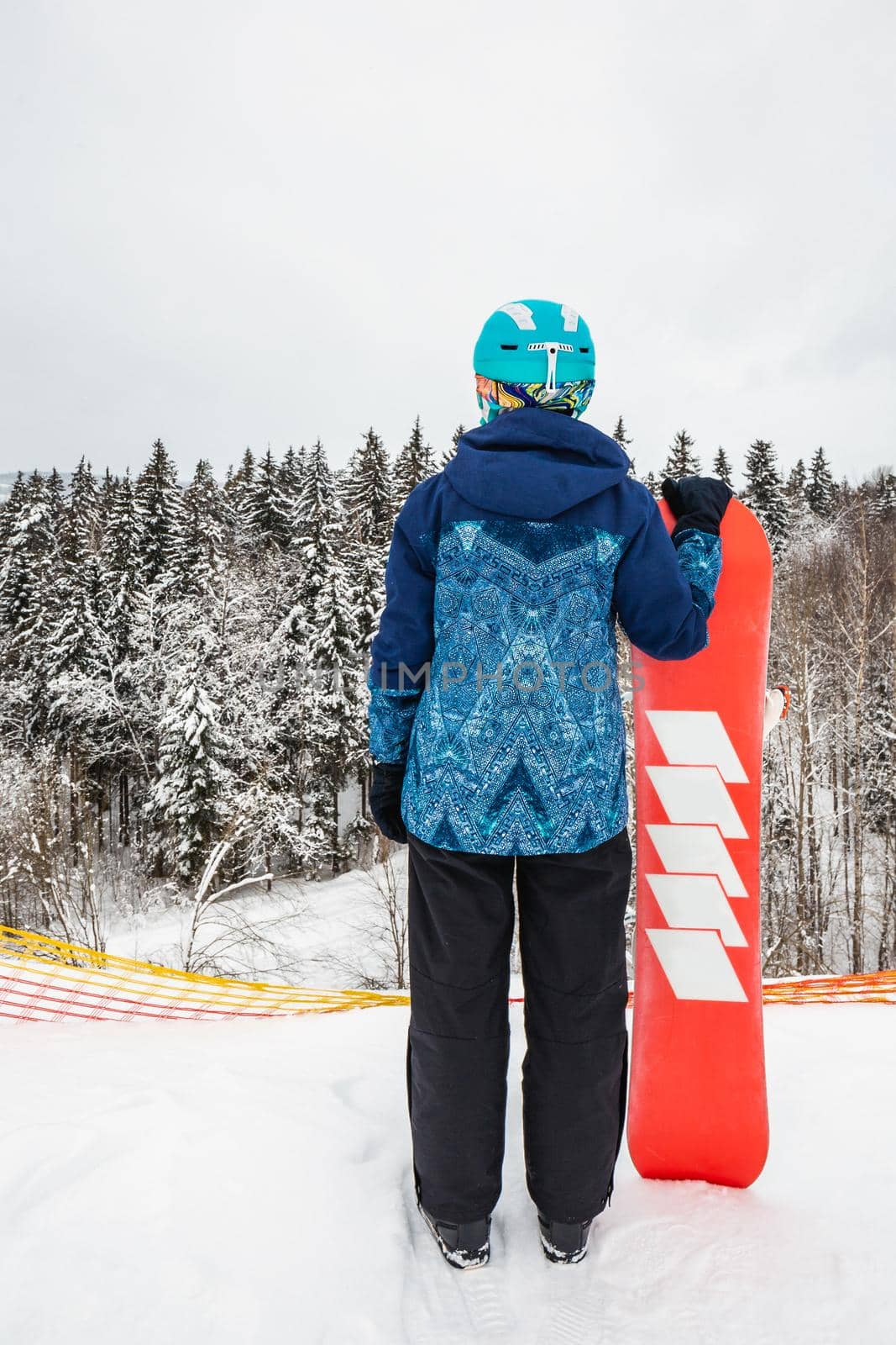 Female in a blue suit standing with a snowboard on a large snow slope. Snowboarder looks at a beautiful view of the snowy forest. Skiing holidays. Logoisk, Belarus