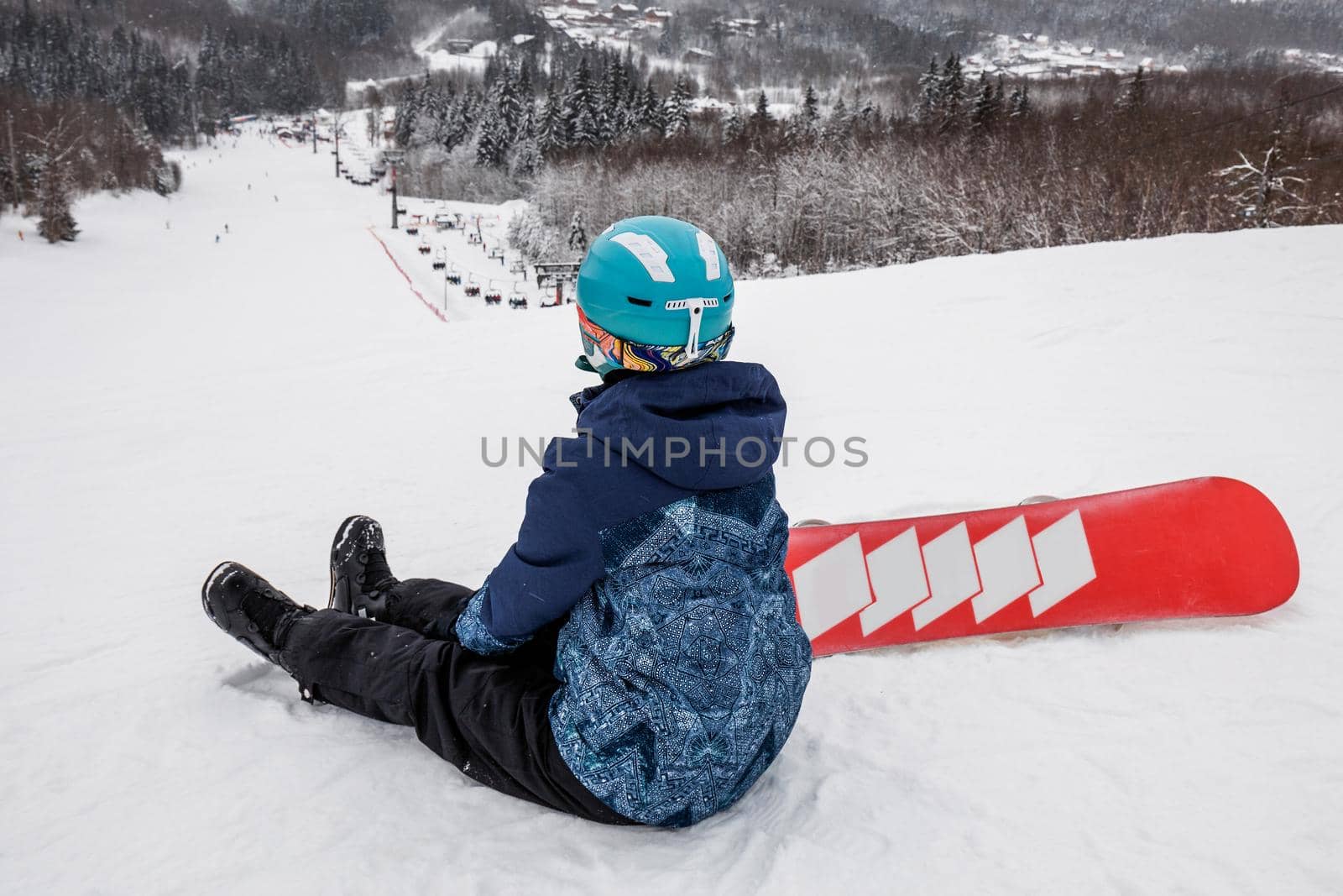 Female in a blue suit sitting with a snowboard on a large snow slope. Snowboarder looks at a beautiful view of the snowy forest. Skiing holidays. Logoisk, Belarus