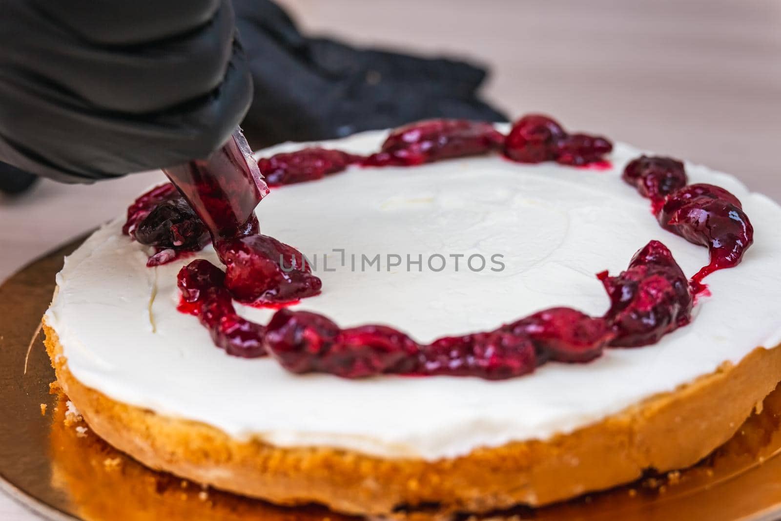Female hand in black glove squeezes out cherry confiture on a cheesecake with coconut.