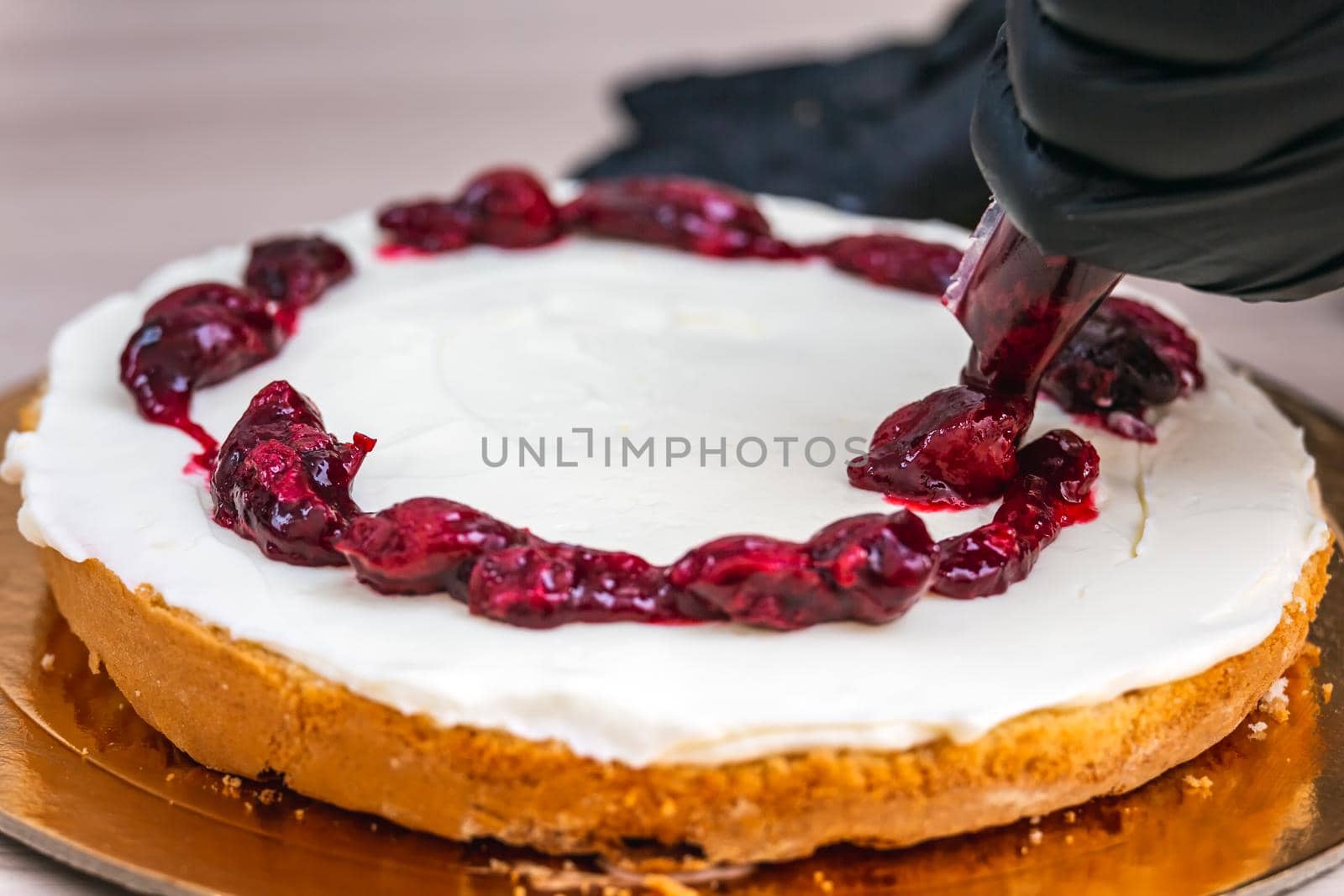 Female hand in black glove squeezes out cherry confiture on a cheesecake with coconut.