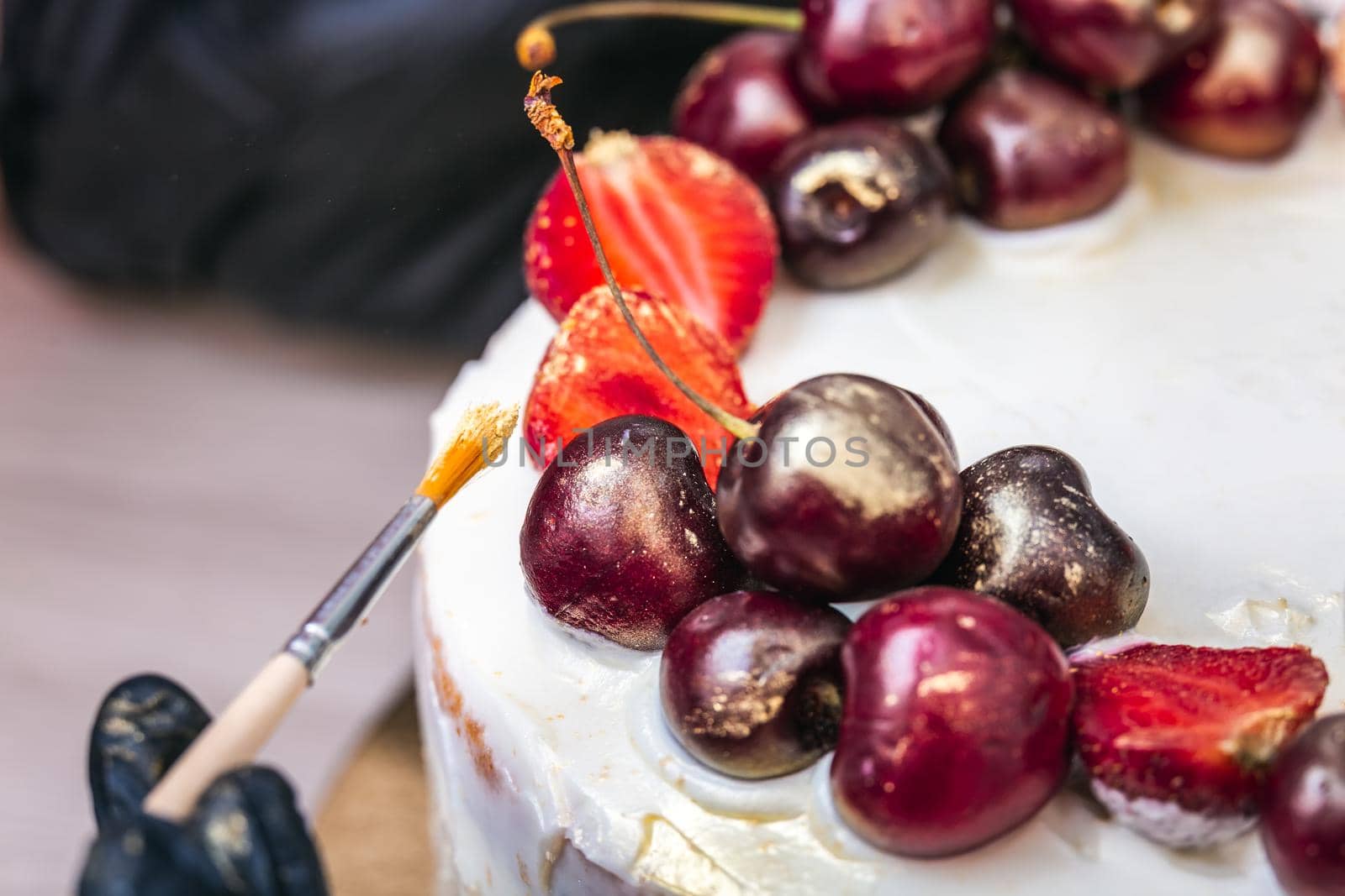 Female hand in black glove puts edible gold on cherries and strawberries on top of cheesecake with coconut.