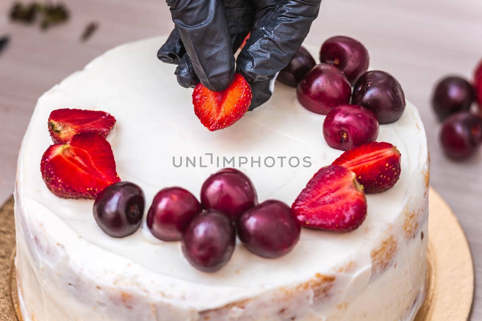 Female hand in black glove puts cherries and strawberries on top of cheesecake with coconut.