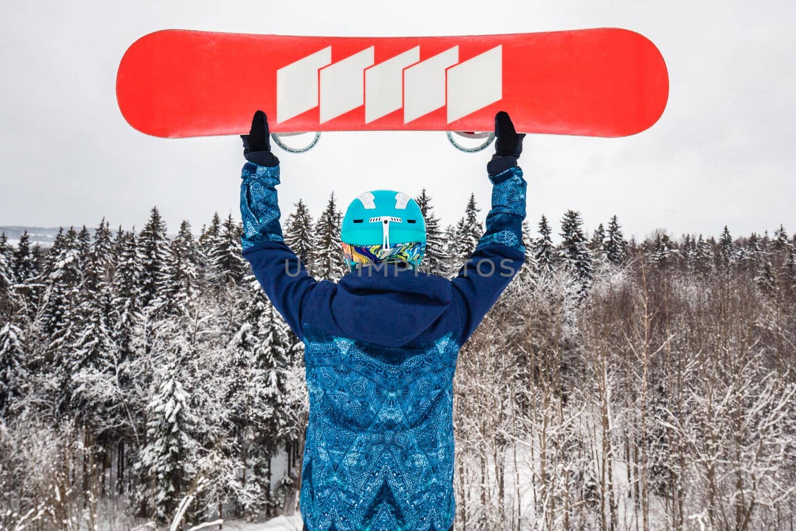 Female in a blue suit standing with a snowboard on a large snow slope. Snowboarder holds the snowboard over his head. Skiing holidays. Logoisk, Belarus