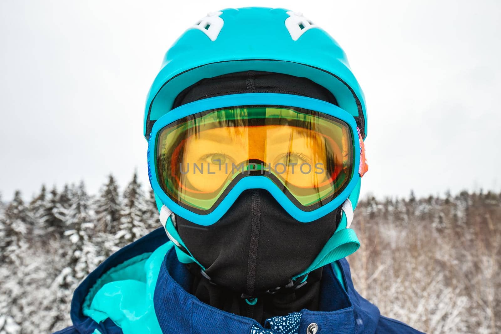 Female in ski mask with blue eyes. Snowboarder looking at camera. Behind there is a beautiful view of the snow-covered forest. Ski vacation. Logoisk, Belarus