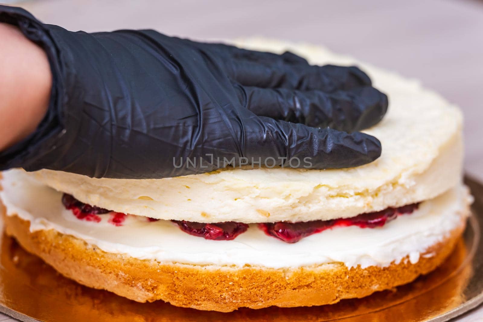 Female hand in black glove puts top layer on a cherry confiture of a coconut cheesecake.