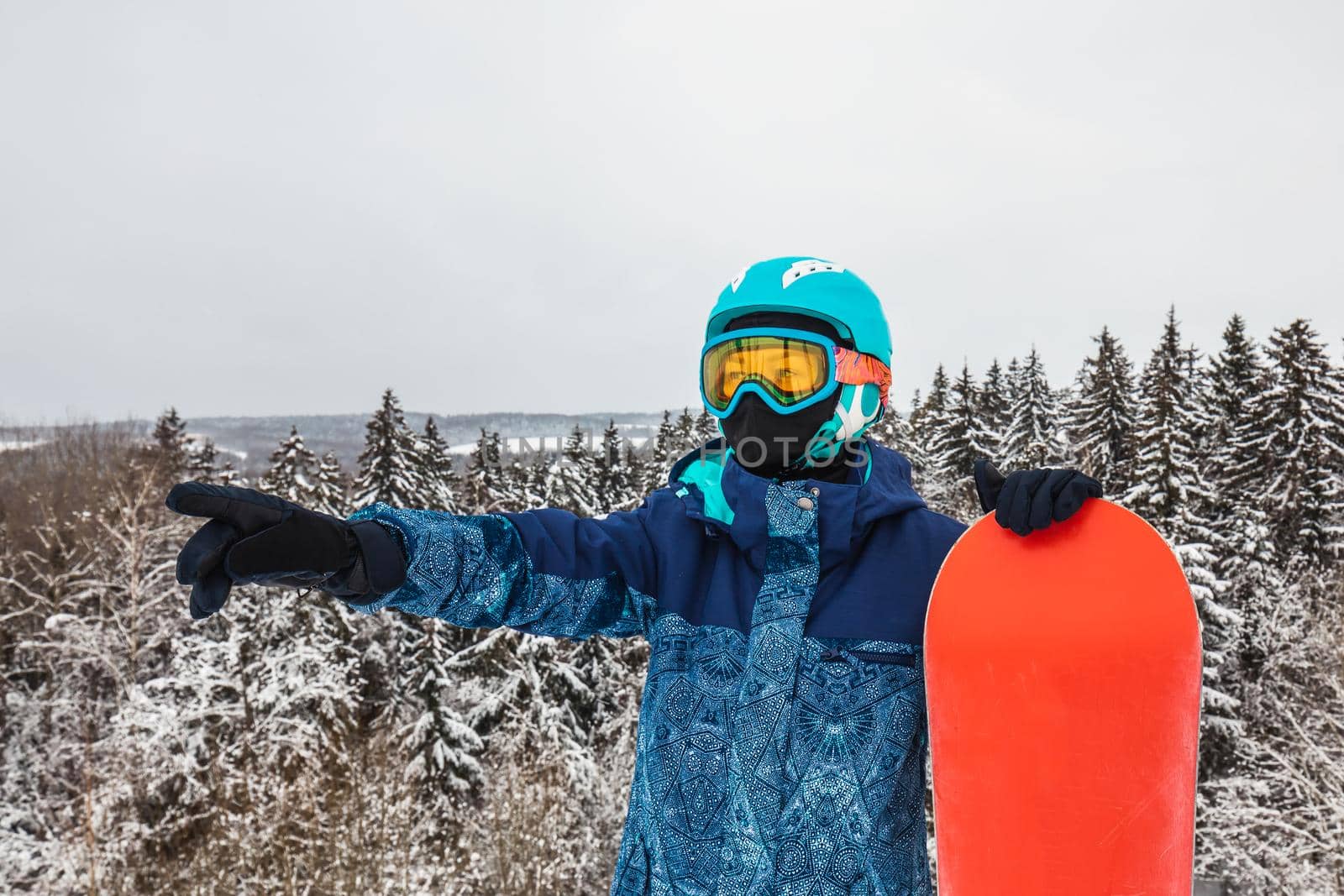 Female in a blue suit standing with a snowboard on a large snow slope. Snowboarder looks at a beautiful view of the snowy forest. Skiing holidays. Logoisk, Belarus