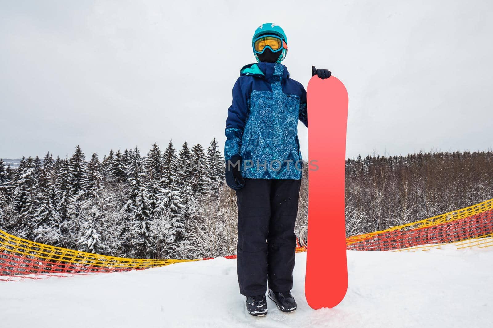 Female in a blue suit standing with a snowboard on a large snow slope. Snowboarder looks at a beautiful view of the snowy forest. Skiing holidays. Logoisk, Belarus