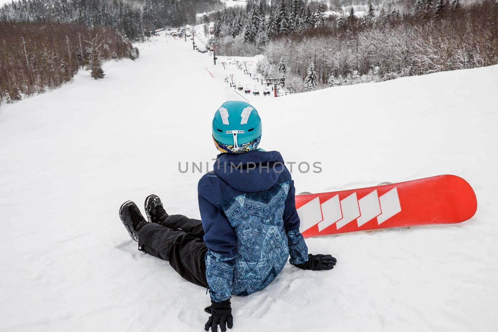Female in a blue suit sitting with a snowboard on a large snow slope. Snowboarder looks at a beautiful view of the snowy forest. Skiing holidays. Logoisk, Belarus