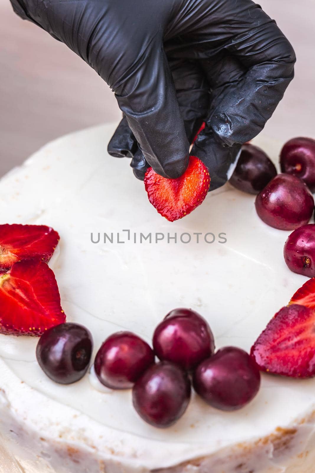 Female hand in black glove puts cherries and strawberries on top of cheesecake with coconut.