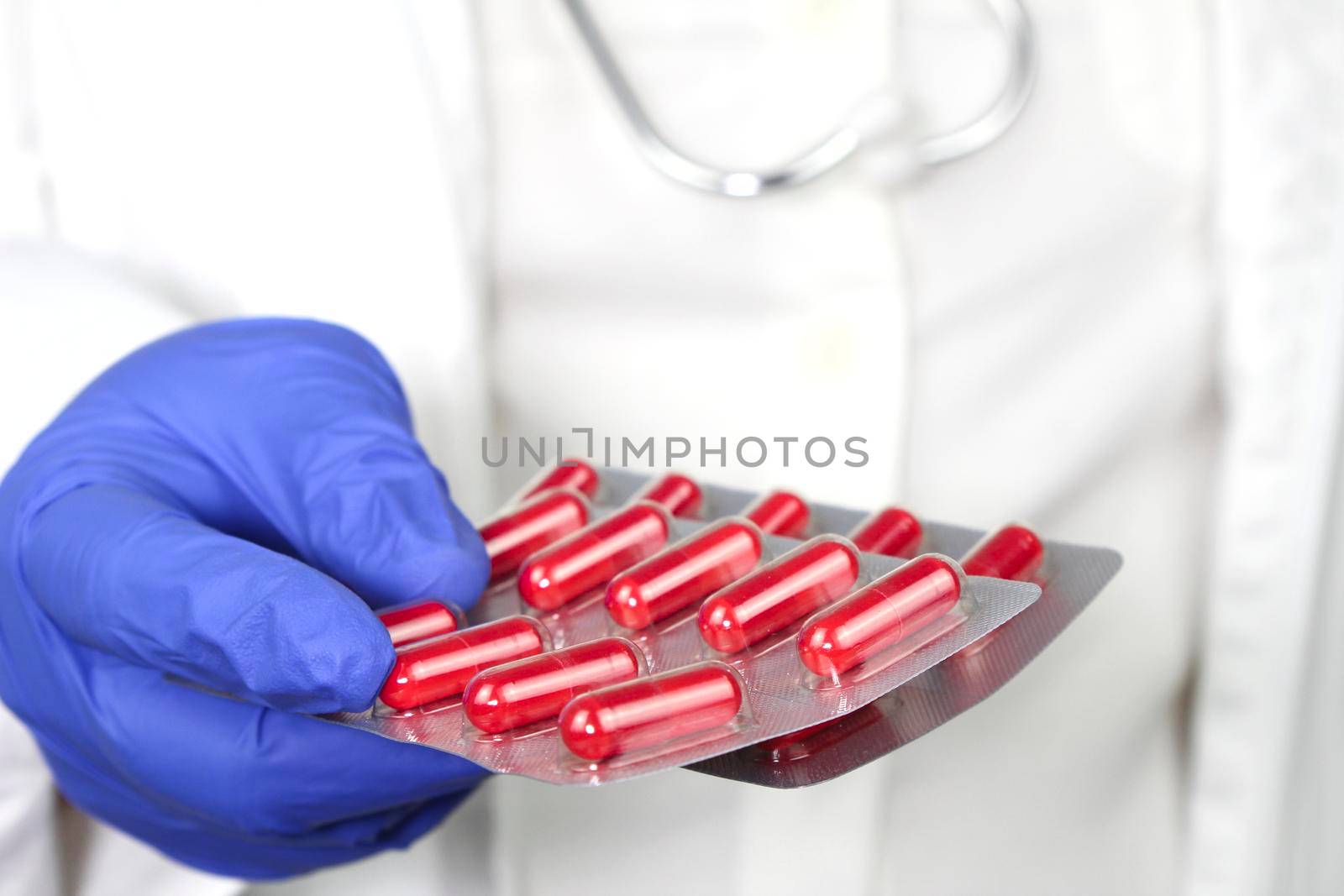 Doctor in blue surgical gloves and wearing stethoscope holding red capsule blisters