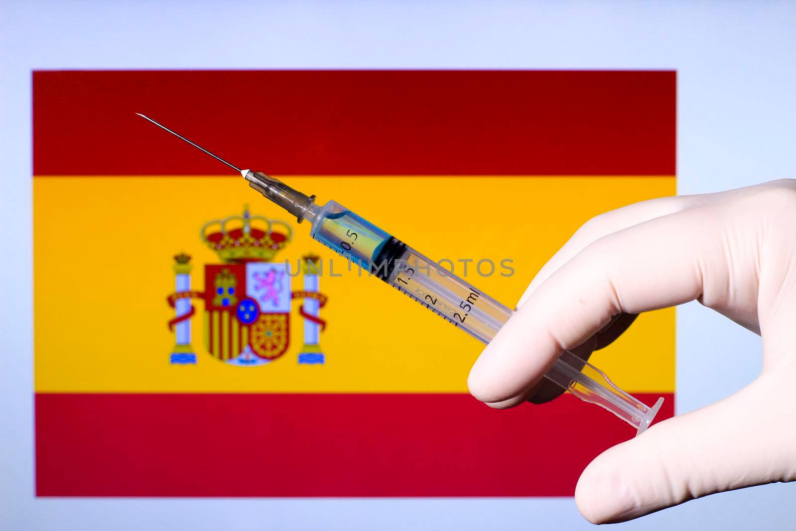 Hand in surgery glove holding syringe with covid vaccine. Spanish flag in the background