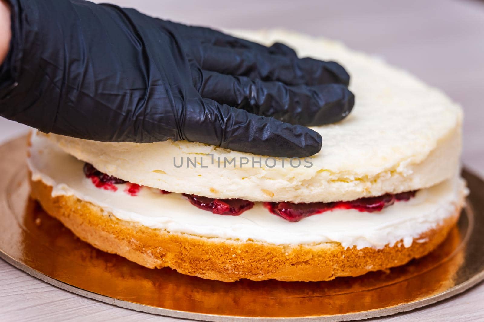 Female hand in black glove puts top layer on a cherry confiture of a coconut cheesecake.