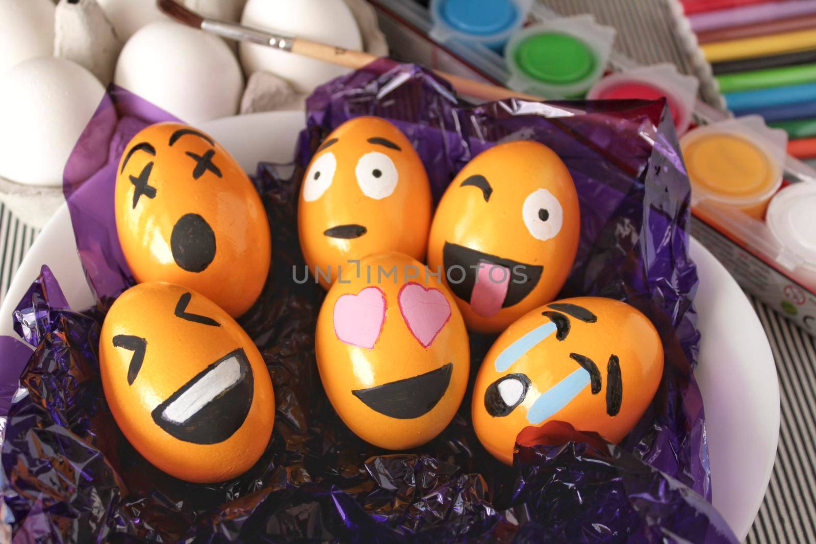 Alicante, Spain- March 18, 2021:Emoticons Easter Eggs on purple cellophane paper. More white eggs, brush and paints in the background.