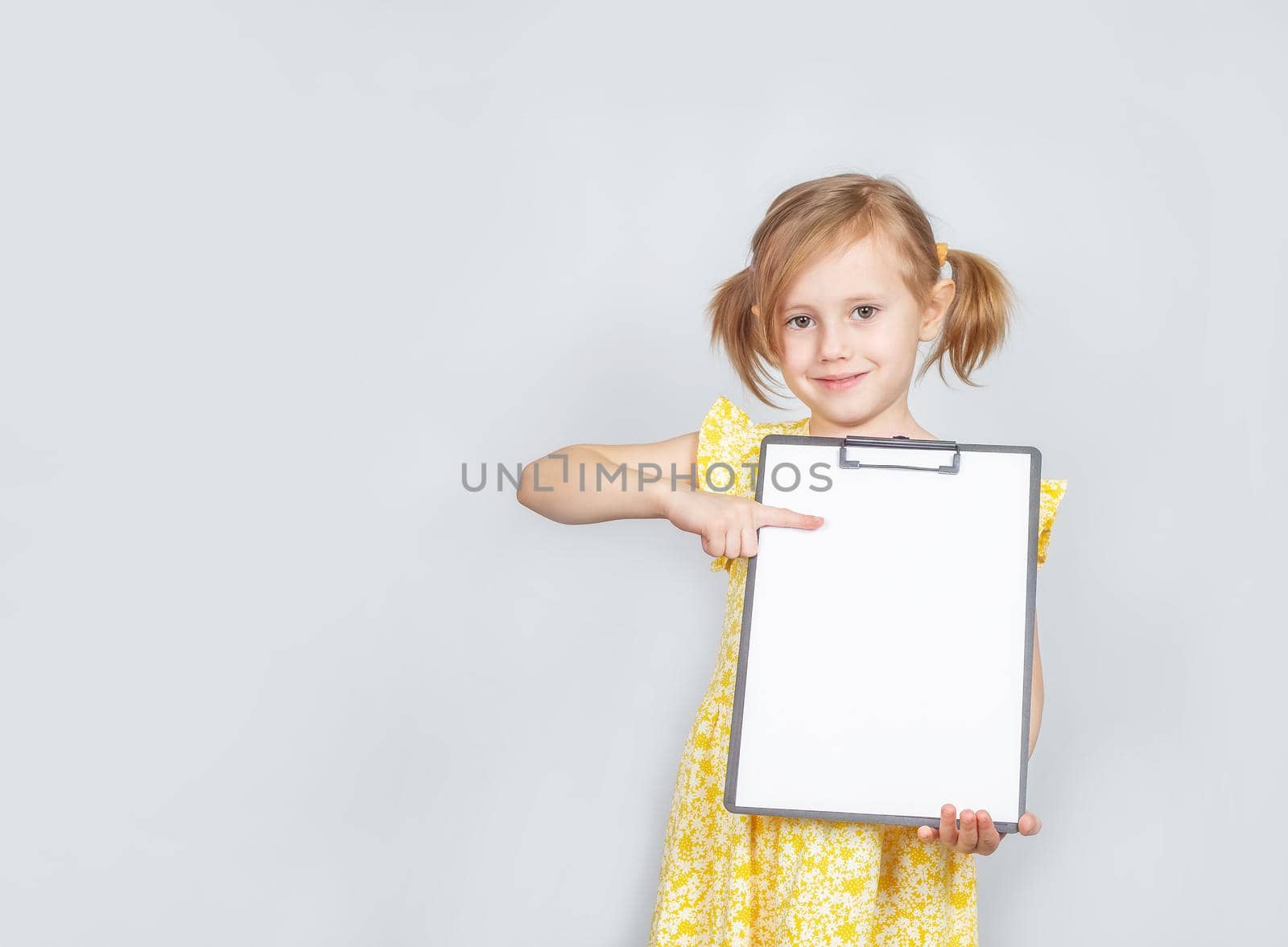 Little Caucasian girl holding a clipboard with a blank paper with copy space for inscription, advertisement posing against studio wall. Advertising concept