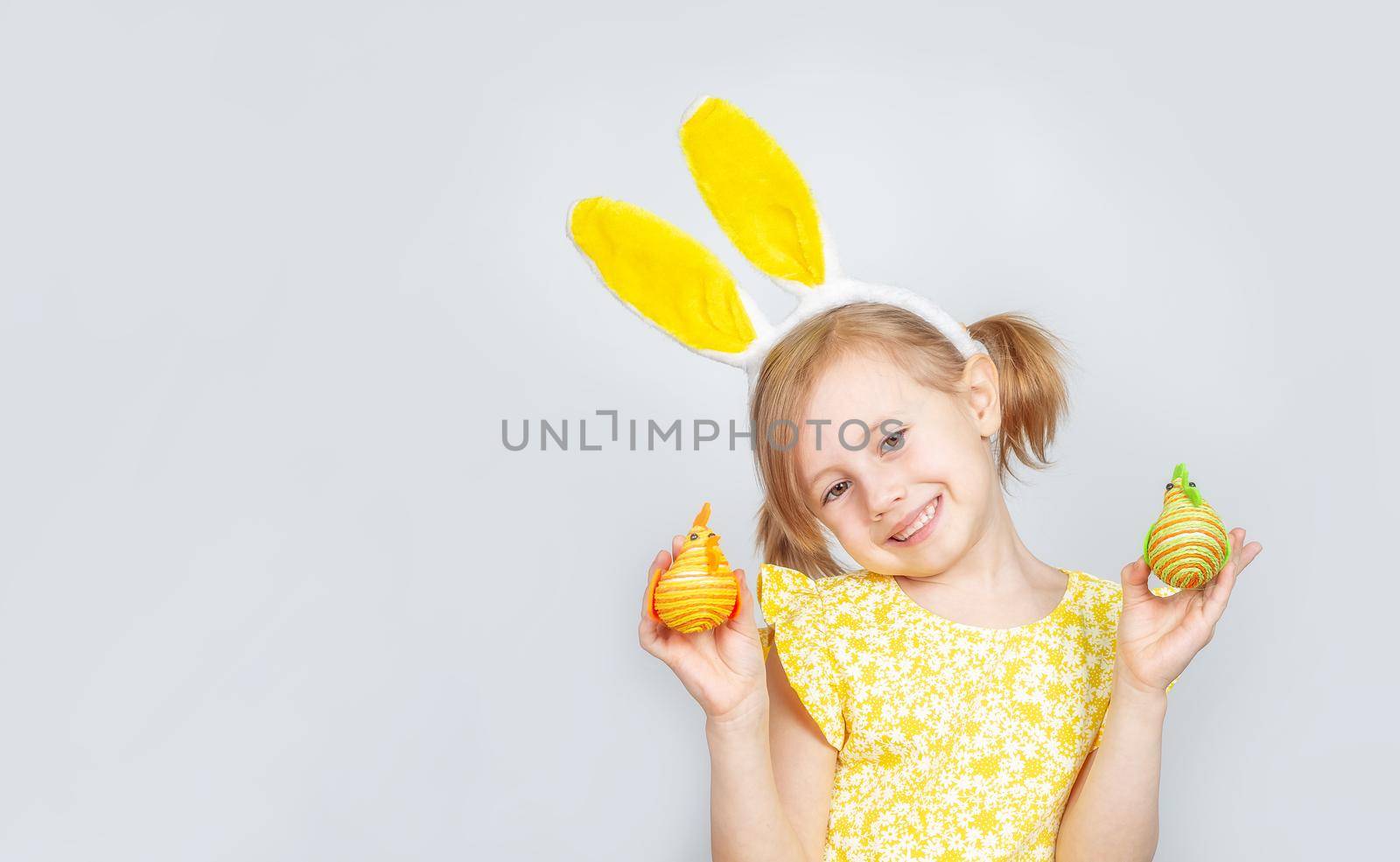 Portrait of a little cute Caucasian smiling girl with bunny ears and decorations for Easter in hands on a gray background. Easter background with place to insert text.