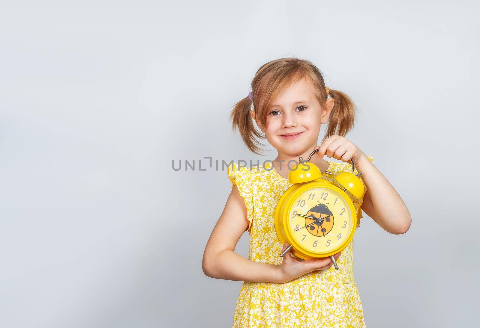 A positive Little Caucasian girl holds an alarm clock in her hand and smiles, advertisement posing against studio wall. Advertising concept