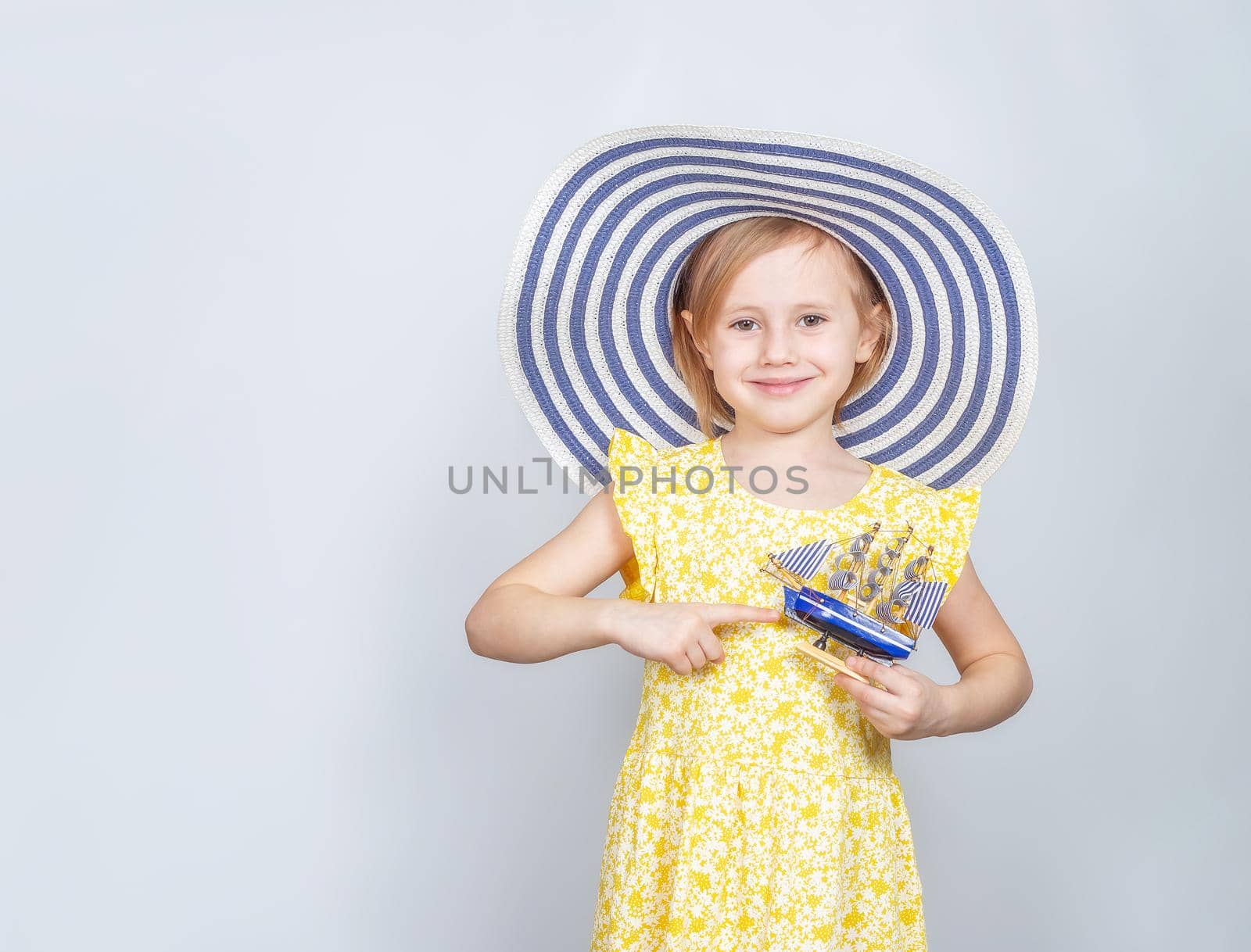 A Little Caucasian in a wide-brimmed hat points her finger at a toy boat. The concept of summer vacation, travel, opening of the beach season.