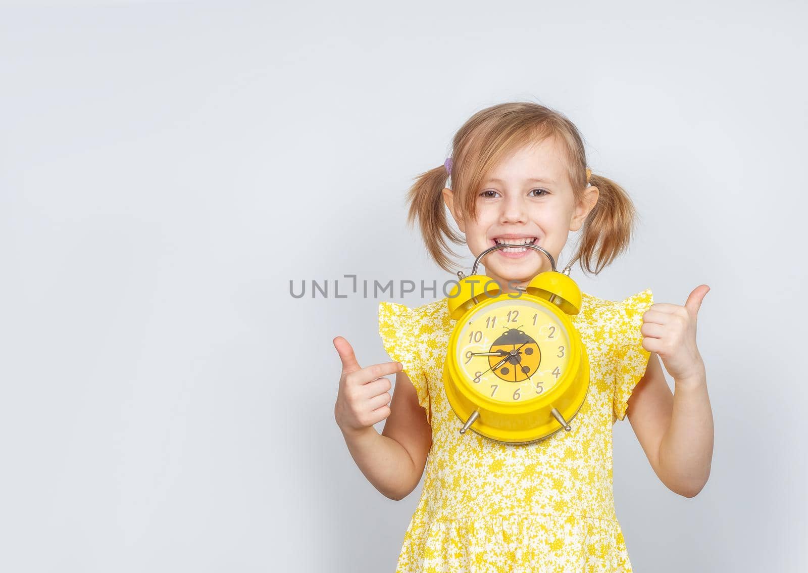 A Little Caucasian girl holds an alarm clock in her teeth and shows two thumbs up, advertisement posing against studio wall. Advertising concept