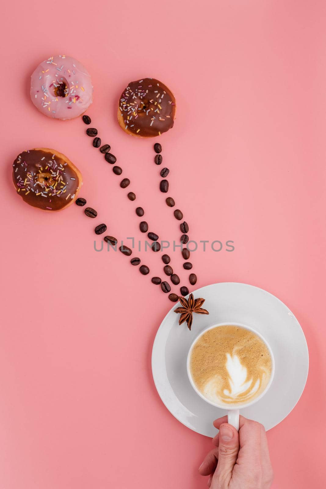 hand holding cup of coffee, beans and three doughnut on pink background