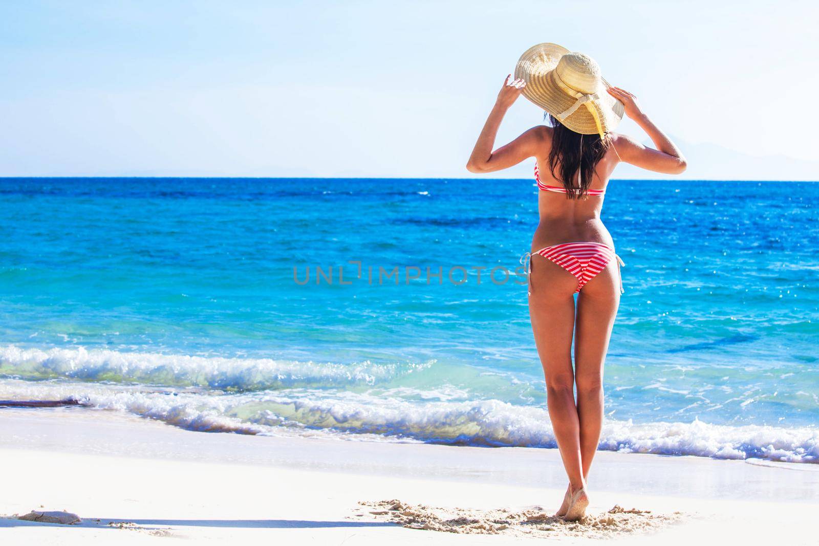 Woman in sunhat and bikini standing on ocean beach on hot summer day