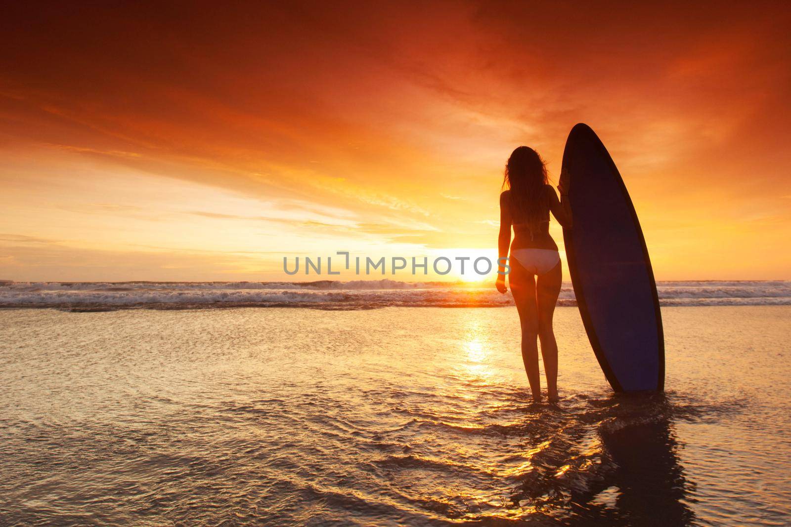 Silhouette of woman on tropical beach holding surfboard at sunset in Bali