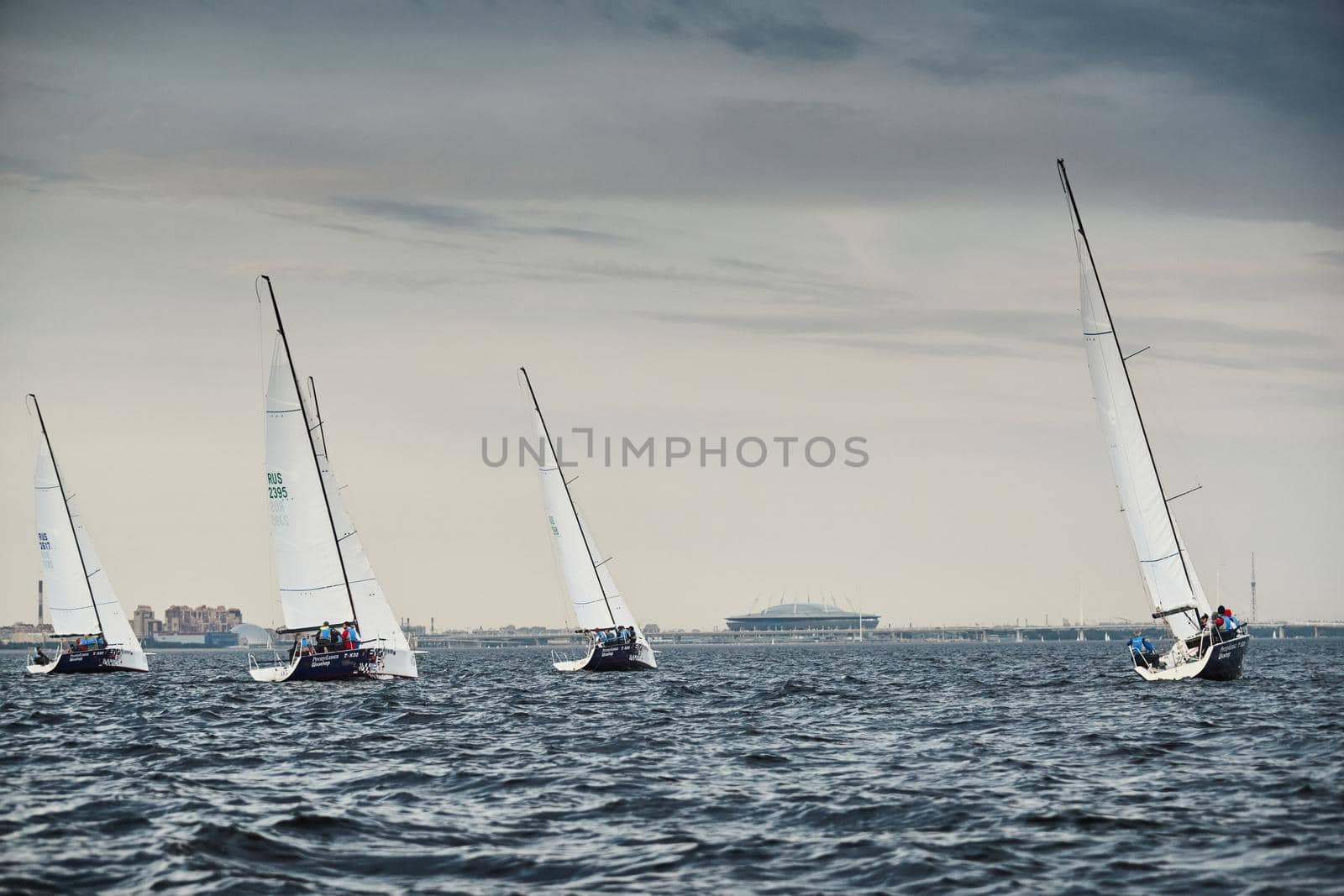 Russia, St.Petersburg, 05 September 2020: Some sailboats in a list goes by sea, the storm sky, regatta, Gazprom arena stadium on background. High quality photo