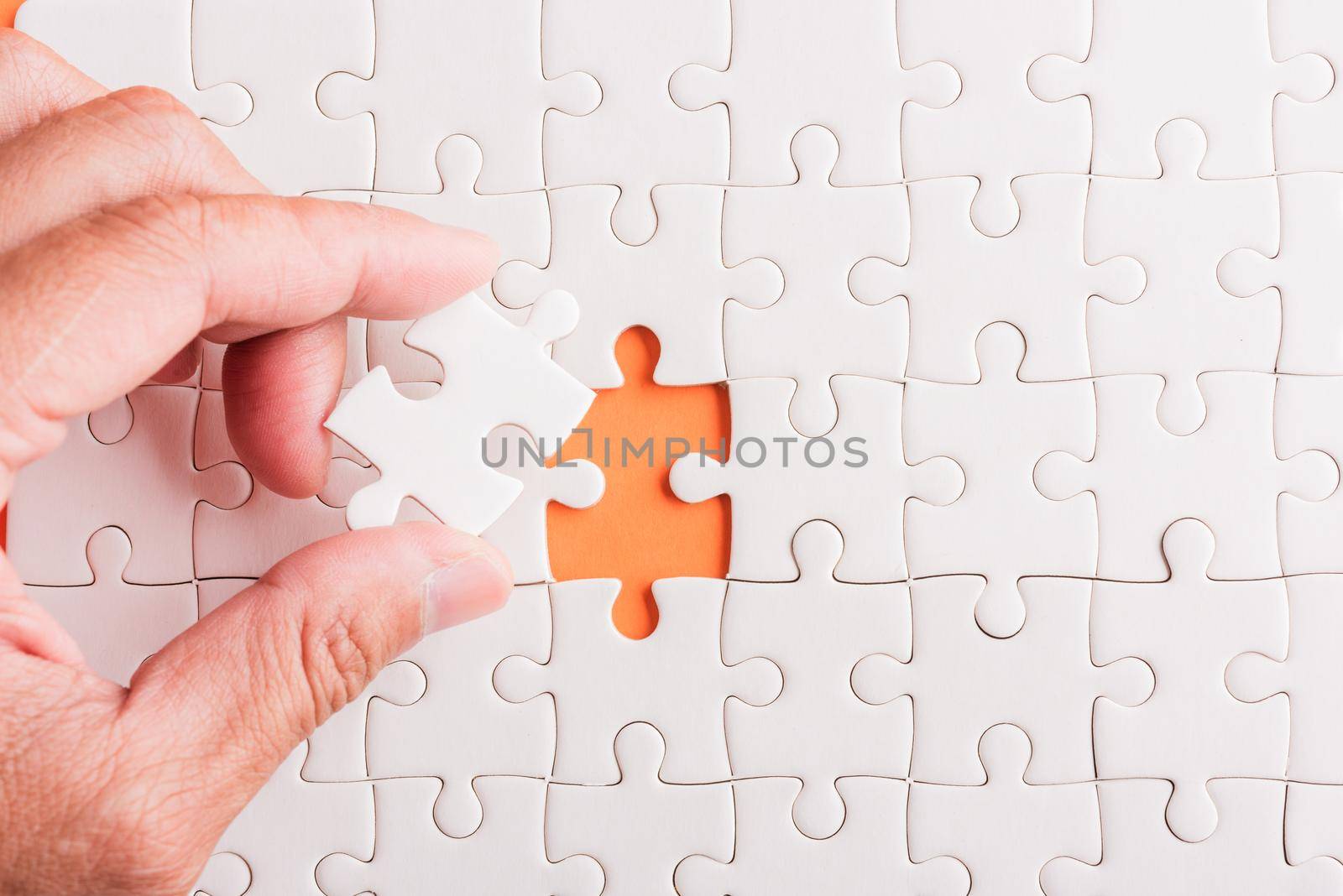 Top view flat lay of hand-holding last piece white paper jigsaw puzzle game last pieces put to place for solve problem complete mission, studio shot on an orange background, quiz calculation concept