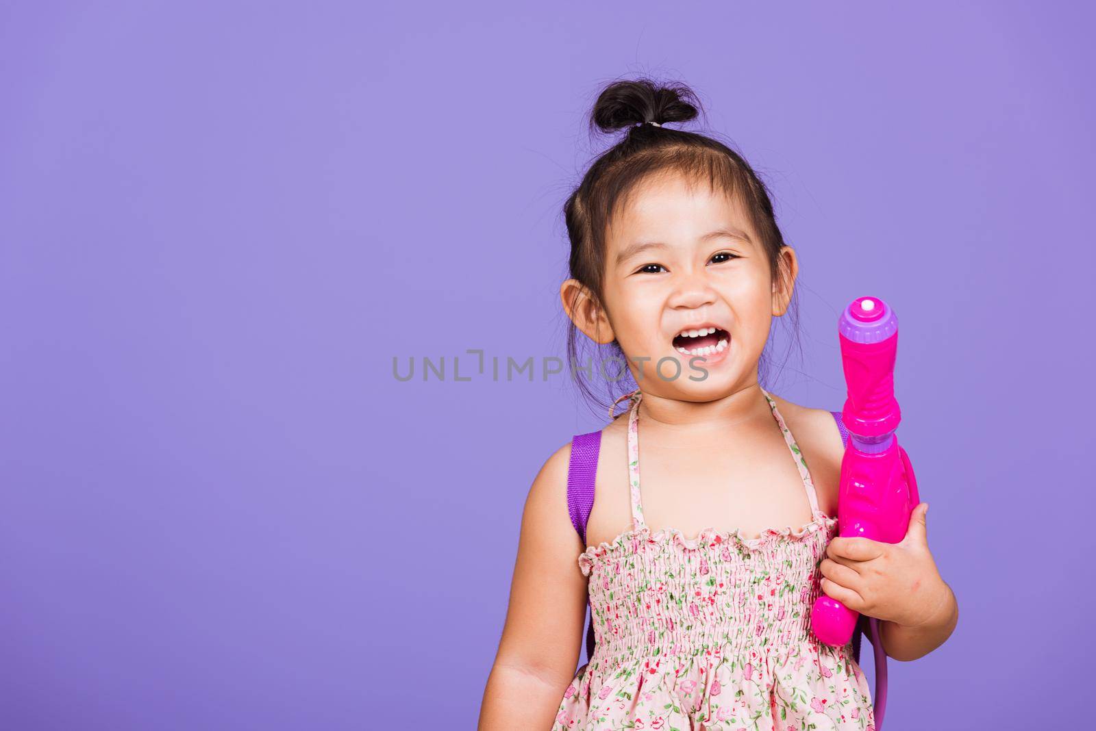 Happy Asian little girl holding plastic water gun, Thai child funny hold toy water pistol and smile, studio shot isolated on purple background, Thailand Songkran festival day national culture concept