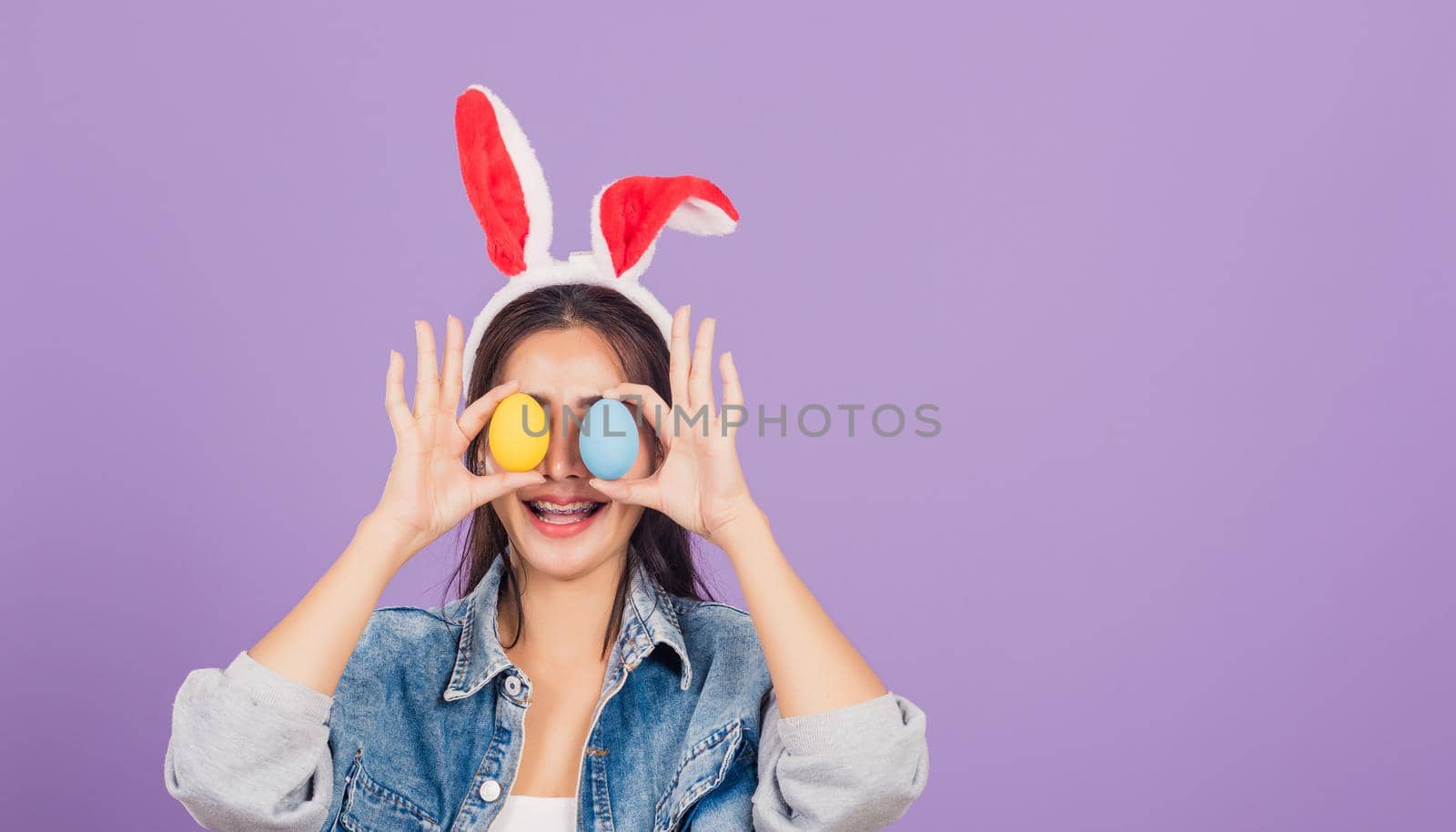 woman smiling wearing rabbit ears and denims holding colorful Easter eggs front eyes by Sorapop
