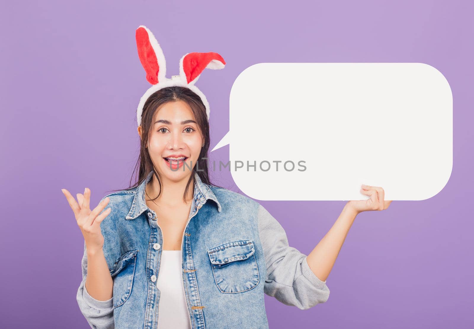 
Happy Easter Day. Beautiful young woman smiling excited wearing rabbit ears and denims holding empty speech bubble, Portrait female looking at camera, studio shot isolated on purple background