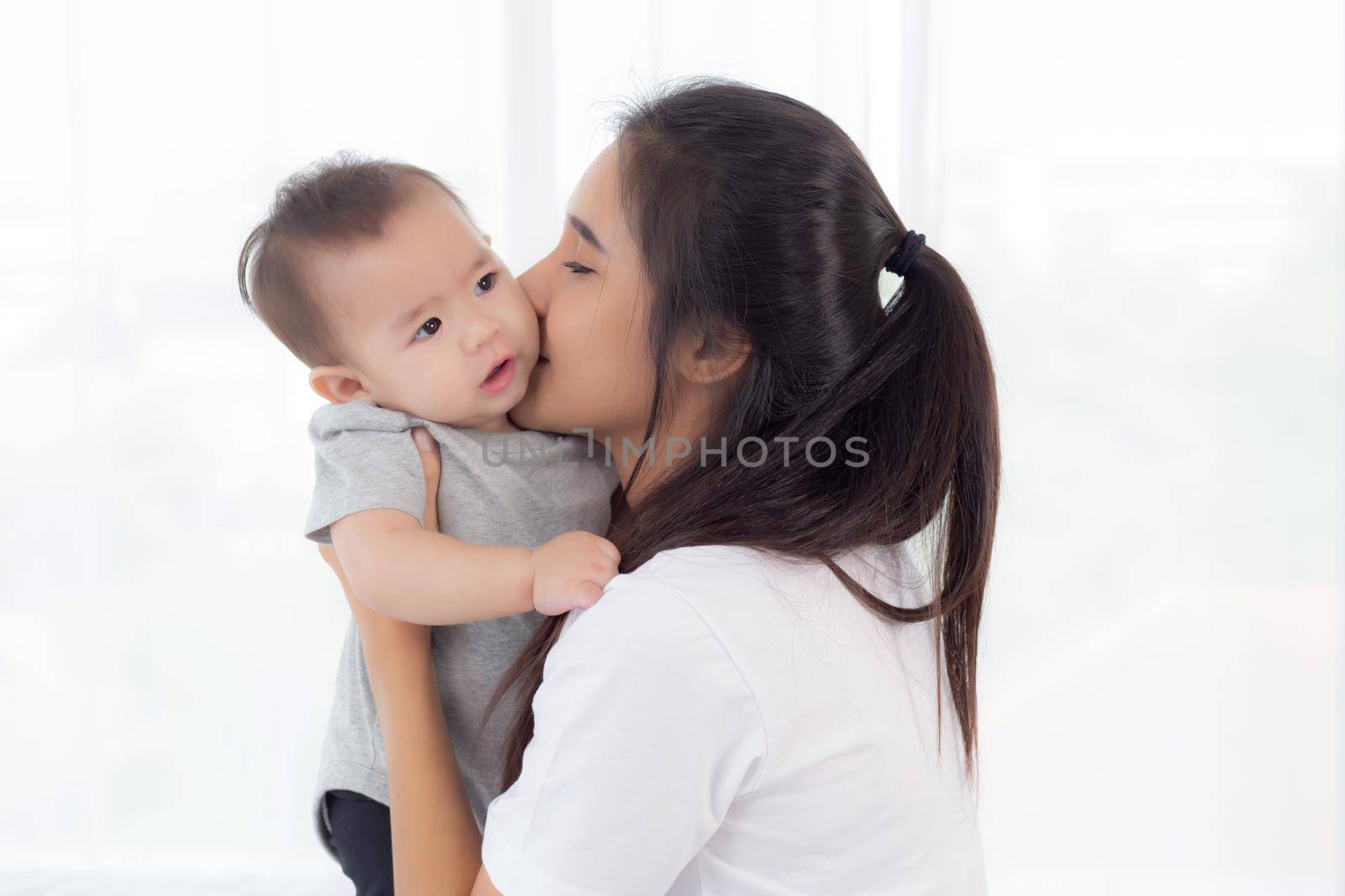 Young asian mother holding little baby girl and kiss together in the bedroom at home, mom carry child and care, woman and kid with carefree, toddler and parent, emotion and expression, family concept.