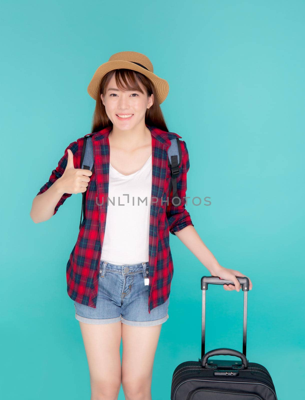 Beautiful young asian woman pulling suitcase isolated on blue background, asia girl cheerful holding luggage walking and gesture thumbs up in vacation with excited, journey and travel concept.