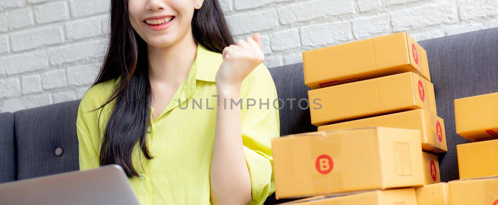 Young asian woman is merchant using laptop computer on sofa checking order of customer with purchase online shopping, business SME or startup, entrepreneur or seller and service delivery product.