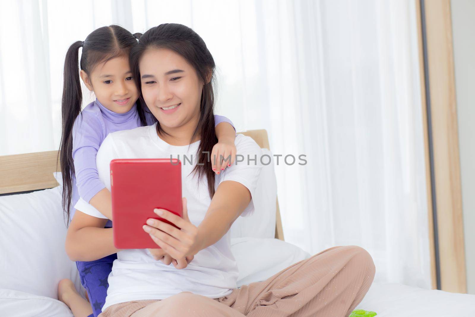 Young asian mom and daughter video call on tablet computer greeting with family together, happy mother and girl with relationship using technology in communication to internet online, two people. by nnudoo