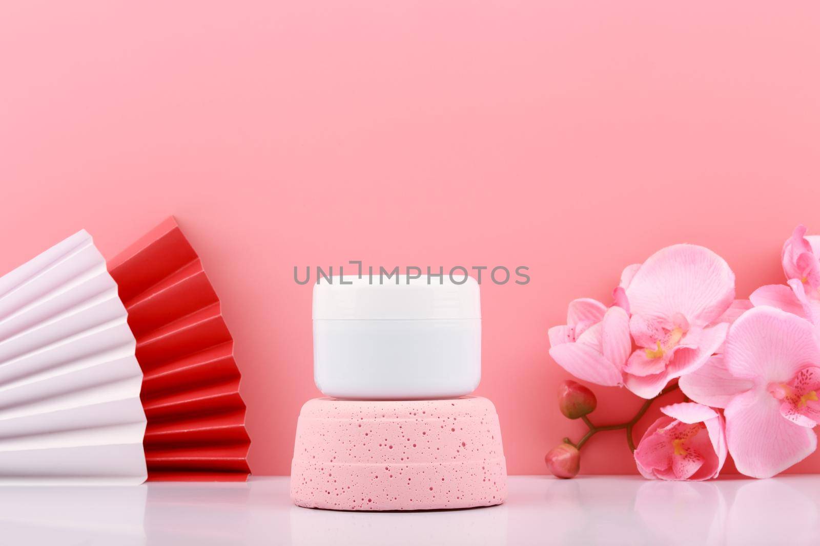 White cosmetic jar on pink pedestal against pink background with copy space decorated with wavers and flowers. Concept of beauty product, hair mask, cream or scrub for skin care.