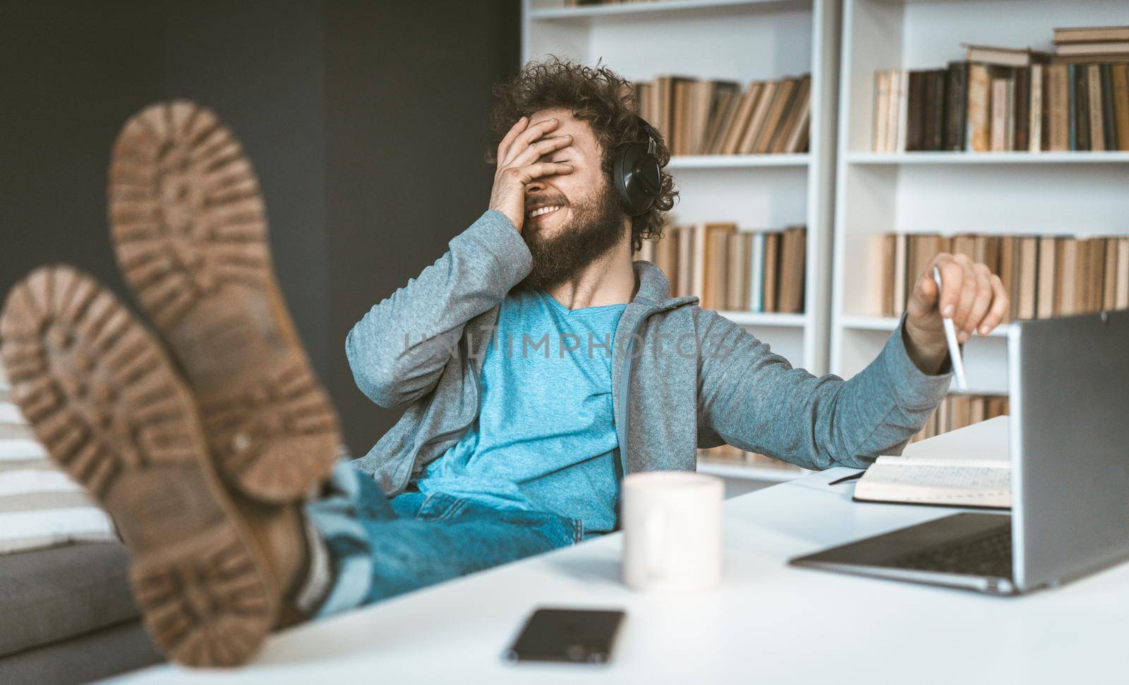 Facepalm. A young programmer works remotely at home. A young man smiles, covering his face with his hands sitting at a table in the office. by LipikStockMedia