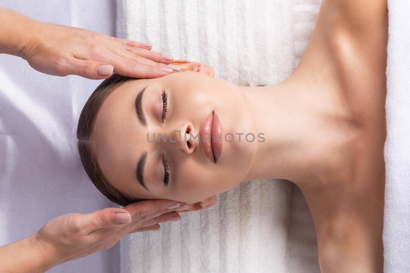 Facial massage, top view of beautiful young woman lying on back and keeping her eyes closed while massage therapist massaging her face